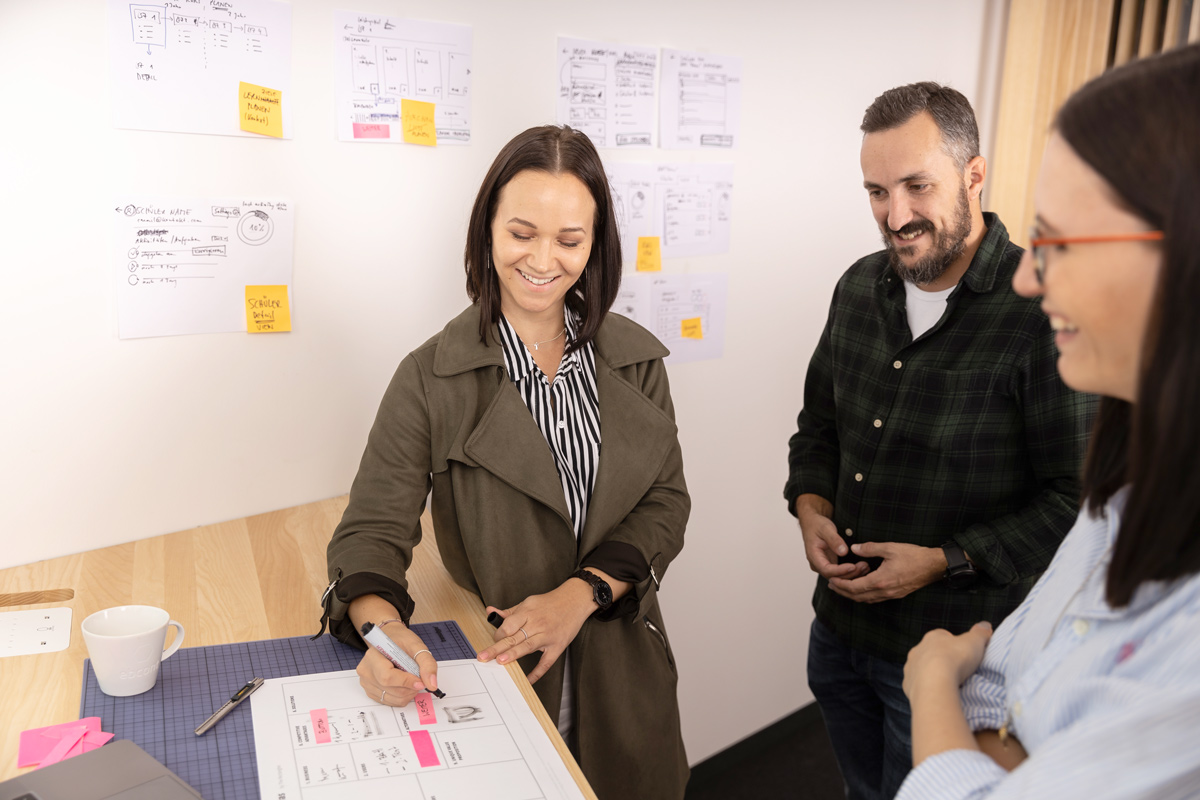 Drei locker gekleidete Kolleg:innen – zwei Frauen und ein Mann – bei einem kreativen Workshop, lächelnd und im Ideenaustausch. Im Hintergrund sind Skizzen und Haftnotizen an einem Whiteboard angebracht.