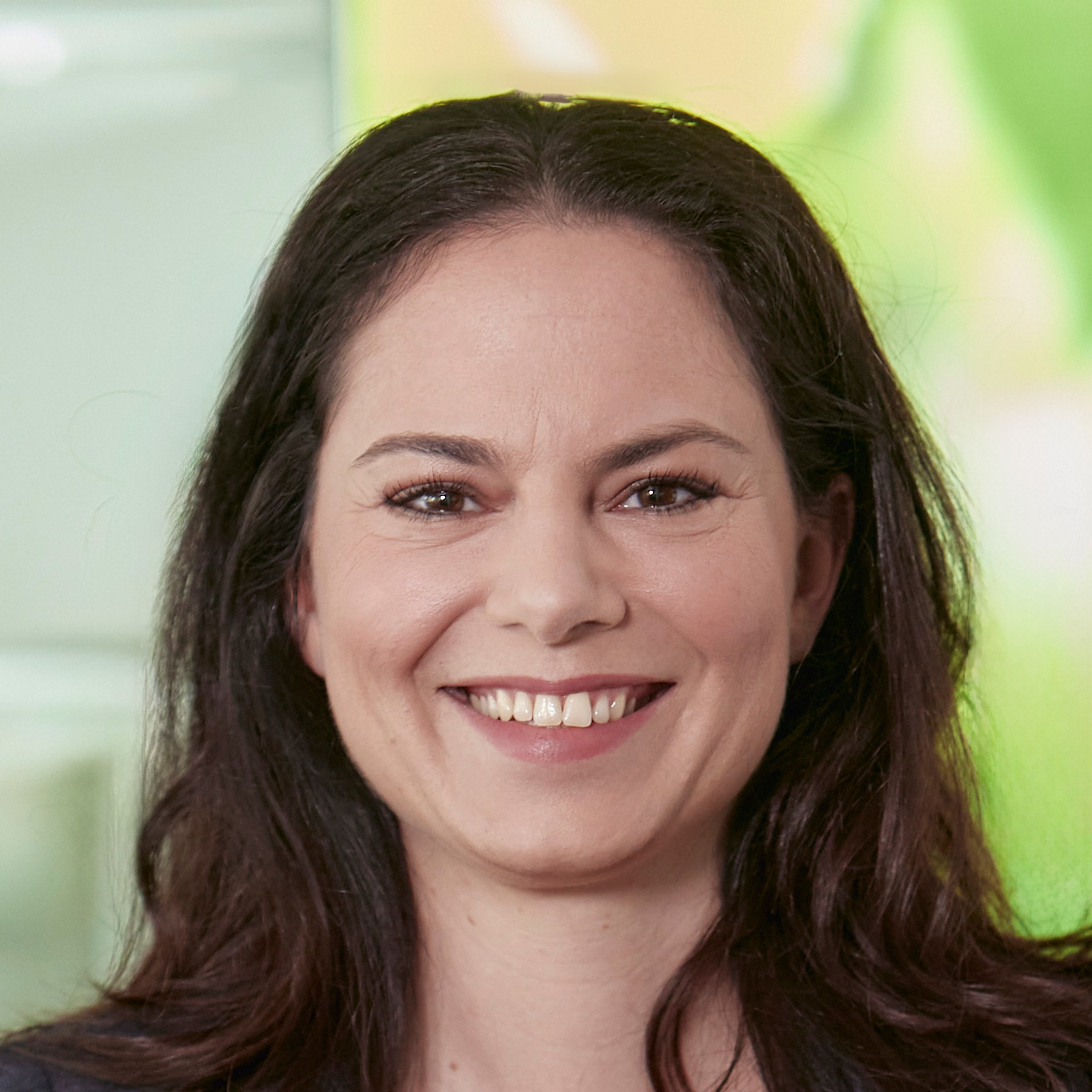 Frau mit langen, dunklen Haaren, lächelt freundlich in die Kamera. Sie trägt ein dunkles Oberteil, im Hintergrund ist eine helle, moderne Umgebung mit grün-gelbem Farbakzent zu erkennen.