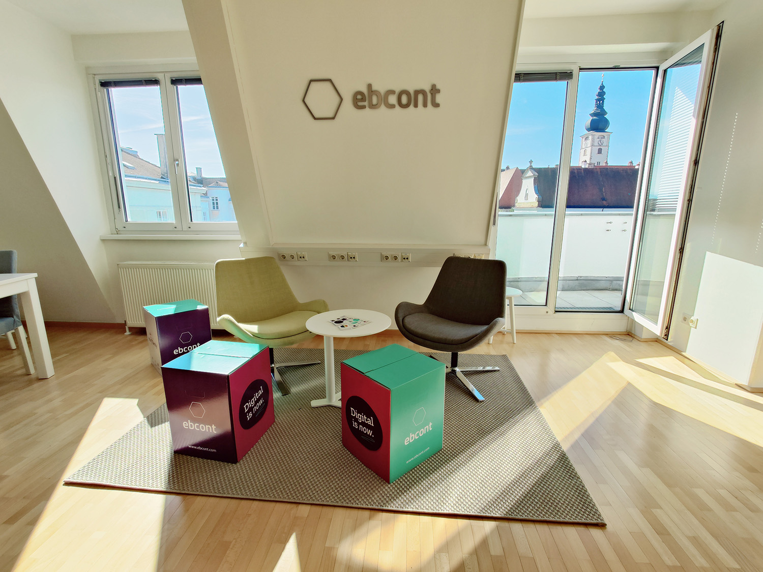 Moderner Besprechungsbereich in einem EBCONT-Büro mit zwei Sesseln, einem kleinen runden Tisch und farbigen Würfeln mit dem Schriftzug „Digital is now.“ sowie „ebcont“. Auf dem Holzboden liegt ein Teppich, im Hintergrund große Fenster mit Blick auf eine Altstadtkirche und blauen Himmel.