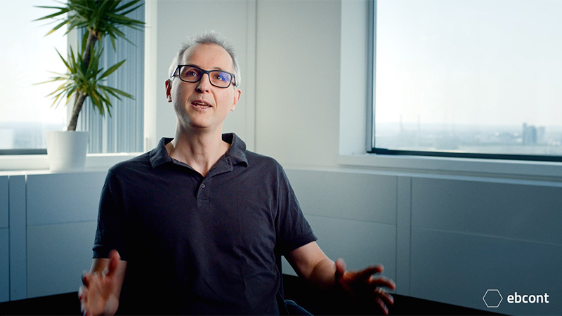 Ausschnitt aus dem Video auf dem Mitarbeiter Thomas entspannt in einem hellen Raum sitzt und lächelt in die Kamera. Er trägt ein schwarzes Polo T-Shirt und Brille.