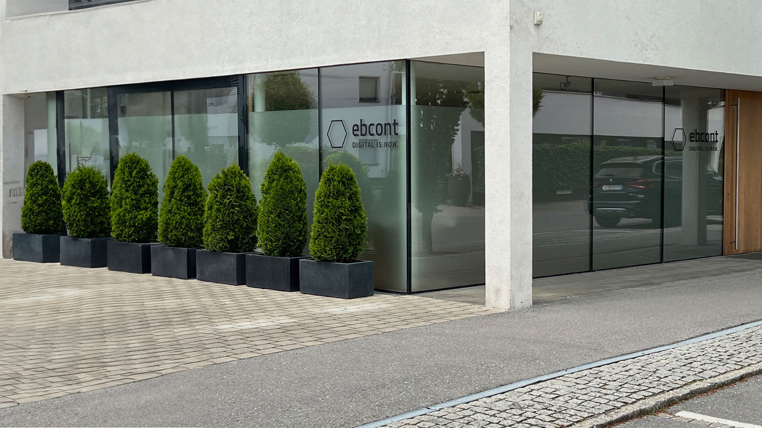 Moderne Glasfassade des EBCONT-Büros in Hard, Vorarlberg. Vor dem Gebäude stehen symmetrisch angeordnete, grüne Zypressen in schwarzen Pflanzgefäßen. Auf den Fenstern ist das EBCONT-Logo mit dem Slogan „DIGITAL IS NOW.“ zu sehen. Rechts führt eine Holztür zum Eingang.