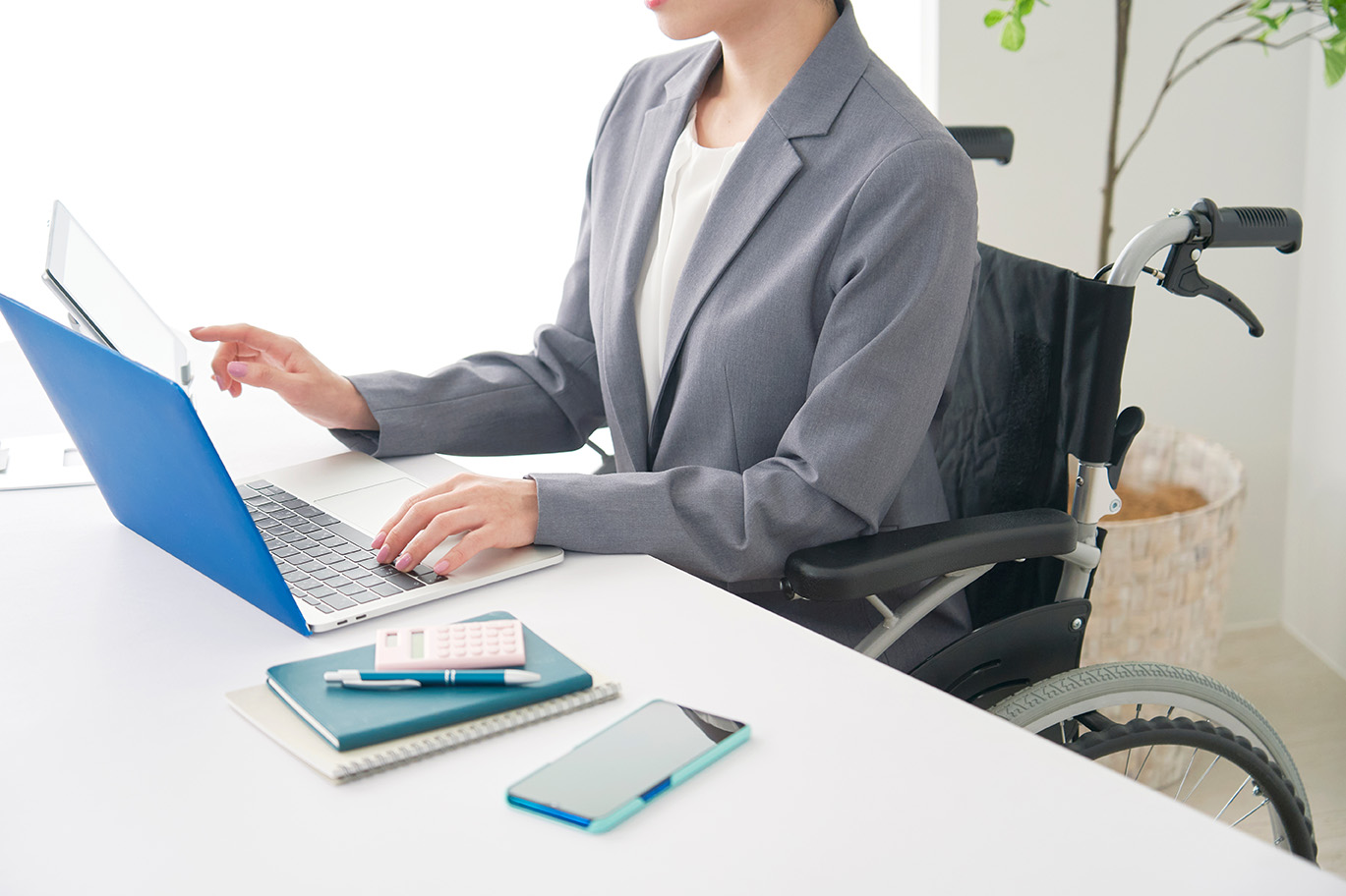 Frau im Rollstuhl arbeitet an einem Laptop mit Tabletaufsatz an einem weißen Schreibtisch, auf dem auch ein Smartphone, Notizbücher, ein Stift und ein Taschenrechner liegen.