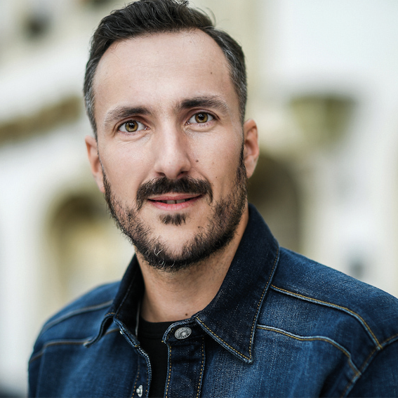 Mann mit dunklem, kurzem Haar und Bart, trägt eine Jeansjacke und blickt direkt in die Kamera. Der Hintergrund ist unscharf, deutet aber auf eine städtische Umgebung mit historischen Gebäudedetails hin.