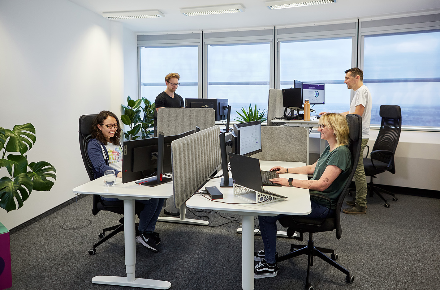 Vier Personen arbeiten in einem modernen EBCONT-Büro an höhenverstellbaren Schreibtischen. Zwei sitzen konzentriert an ihren Laptops, zwei stehen an Monitoren. Große Fenster und Pflanzen sorgen für eine helle, freundliche Atmosphäre.