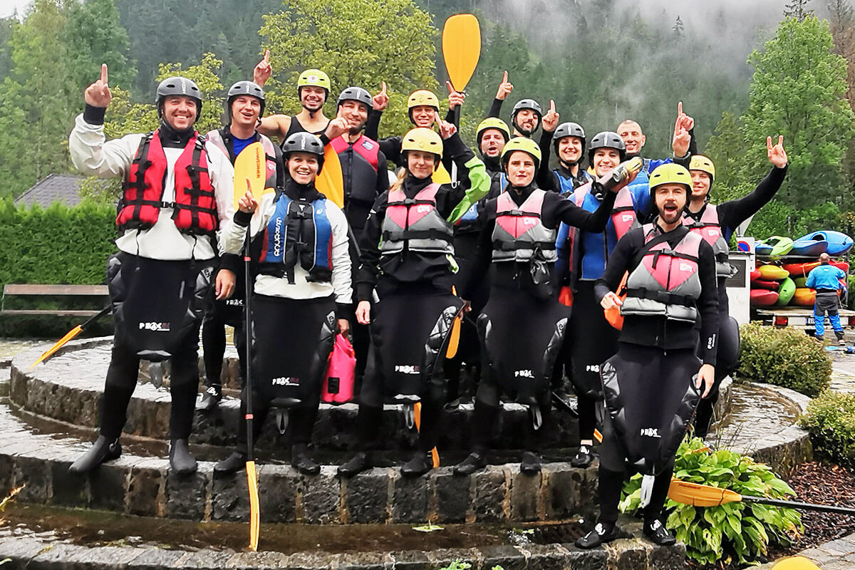 Eine Gruppe von EBCONT-Mitarbeitenden in Neoprenanzügen, Schwimmwesten und Helmen posiert lachend mit Paddeln bei einem Teamevent im Grünen – bereit zum Rafting-Abenteuer.