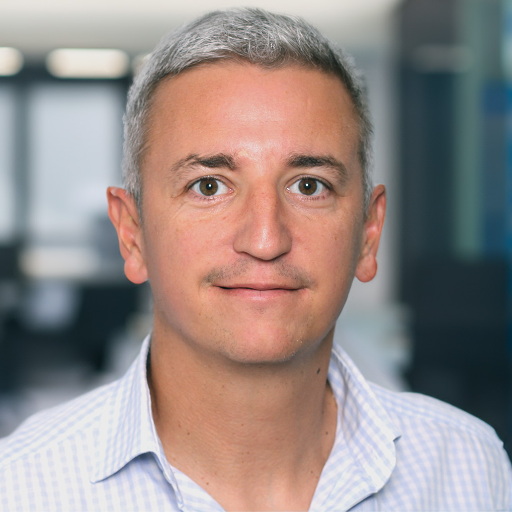 Mann mit kurzen, grauen Haaren und hellem Teint in einem karierten Hemd blickt freundlich in die Kamera. Der Hintergrund ist unscharf und zeigt eine moderne Büro-Umgebung mit Glaswänden und Arbeitsplätzen.