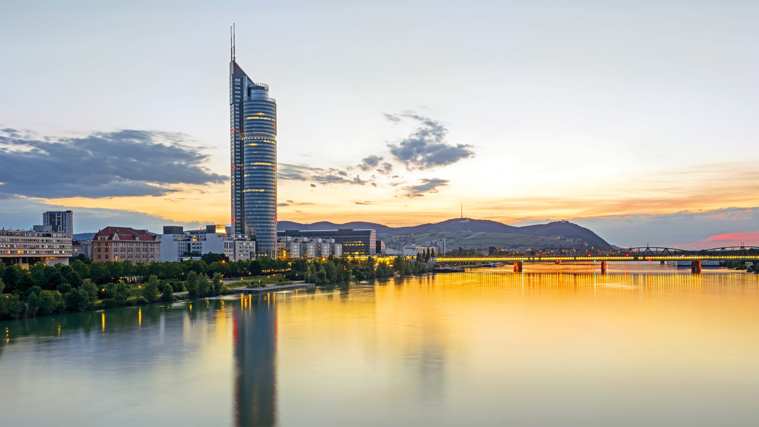 Abendliche Skyline von Wien mit Blick auf den Millennium Tower am Ufer der Donau. Der hohe, runde Glasturm ragt markant in den Himmel, während sich das Licht des Sonnenuntergangs im Wasser spiegelt. Rechts ist eine beleuchtete Brücke zu sehen, im Hintergrund die Hügel des Wienerwalds.