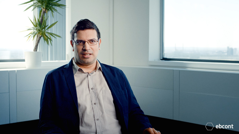 Ausschnitt aus dem Video auf dem Mitarbeiter Frederick entspannt in einem hellen Raum sitzt. Er trägt ein helles Hemd und ein schwarzes Sakko und hat eine Brille.