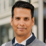 Porträt eines Mannes mit dunklen Haaren im Anzug mit Weste und Krawatte, vor einem unscharfen Hintergrund im Freien.