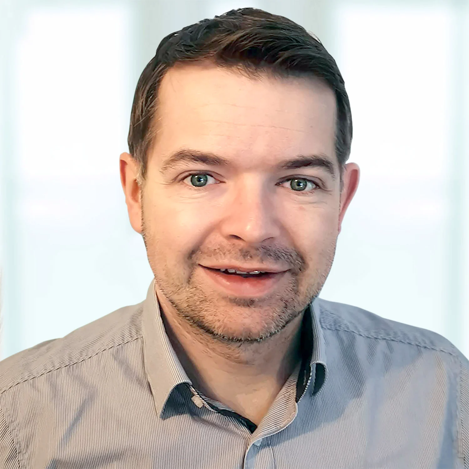 Mann mit kurzen braunen Haaren, leichtem Bart und grün-blauen Augen, trägt ein graues Hemd und blickt lächelnd in die Kamera. Der Hintergrund ist hell und unscharf.