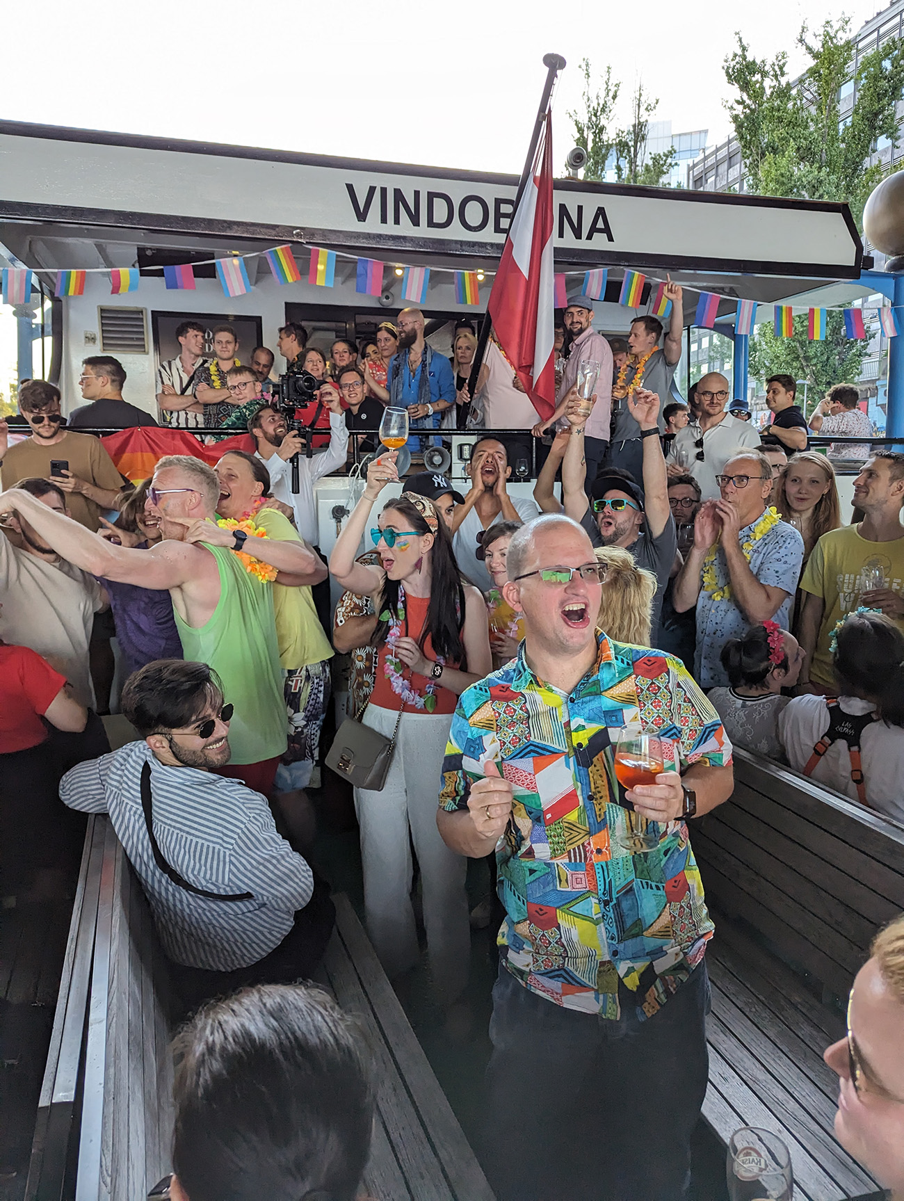 Zahlreiche Menschen feiern ausgelassen an Bord des Schiffs „Vindobona“ beim EBCONT Pride-Boot Event. Regenbogenfahnen und bunte Kleidung sorgen für eine fröhliche Stimmung. Eine österreichische Flagge weht am Heck des Bootes.
