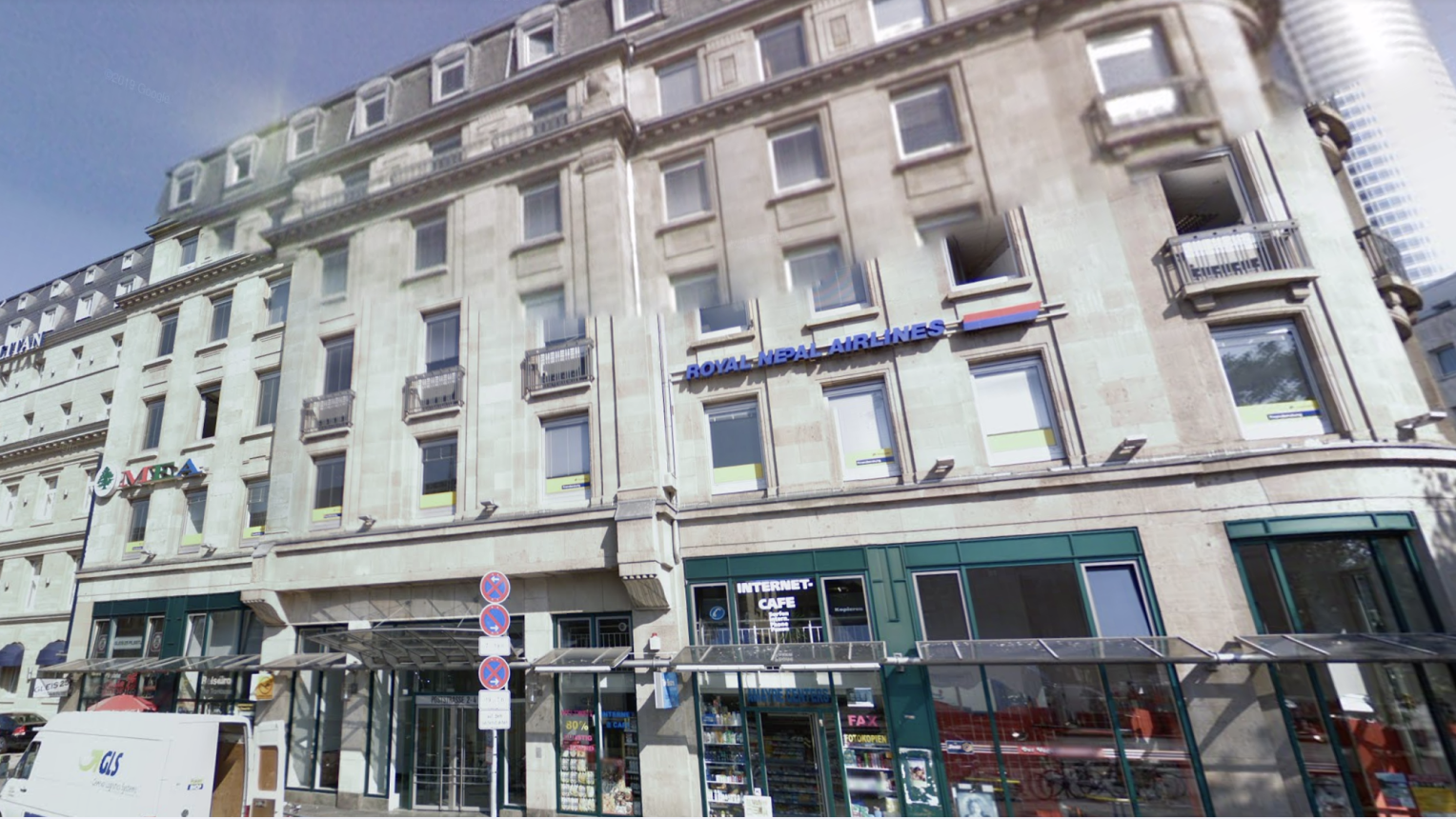 Straßenszene in Frankfurt mit einem mehrstöckigen, historischen Gebäude aus hellem Stein. Im Erdgeschoss befinden sich mehrere Geschäfte, darunter ein Internet-Café und ein Lebensmittelmarkt. An der Fassade im oberen Bereich ist das Schild „Royal Nepal Airlines“ angebracht. Auf dem Gehsteig sind ein Lieferwagen und Verkehrsschilder zu sehen. Im Hintergrund ragt ein moderner Glasturm in den Himmel.
