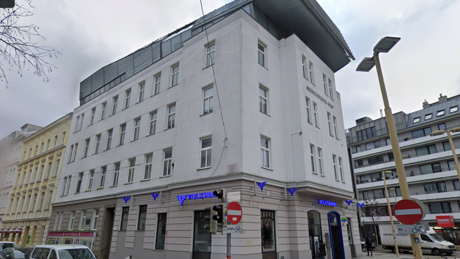 Straßenecke in einer Stadt mit einem fünfstöckigen, weißen Eckgebäude namens „Heßlinger-Haus“. Im Erdgeschoss befindet sich eine Filiale der Volksbank mit blauer Leuchtschrift. Umgeben ist das Gebäude von klassischen Altbauten und modernen Wohnhäusern. Verkehrsschilder, Straßenlaternen und geparkte Fahrzeuge prägen das Straßenbild.