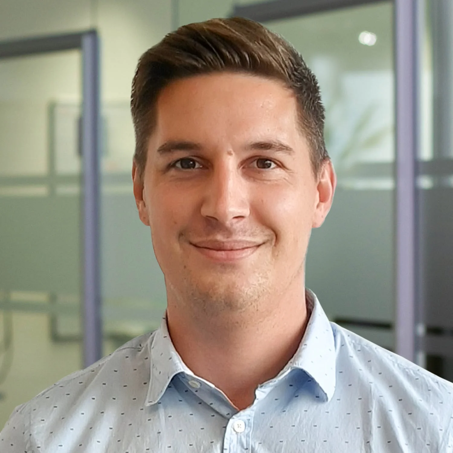 Junger Mann mit kurzen, nach hinten frisierten Haaren trägt ein hellblaues Hemd mit feinem Muster und lächelt in die Kamera. Im Hintergrund ist ein modernes Büro mit Glastrennwänden zu sehen.