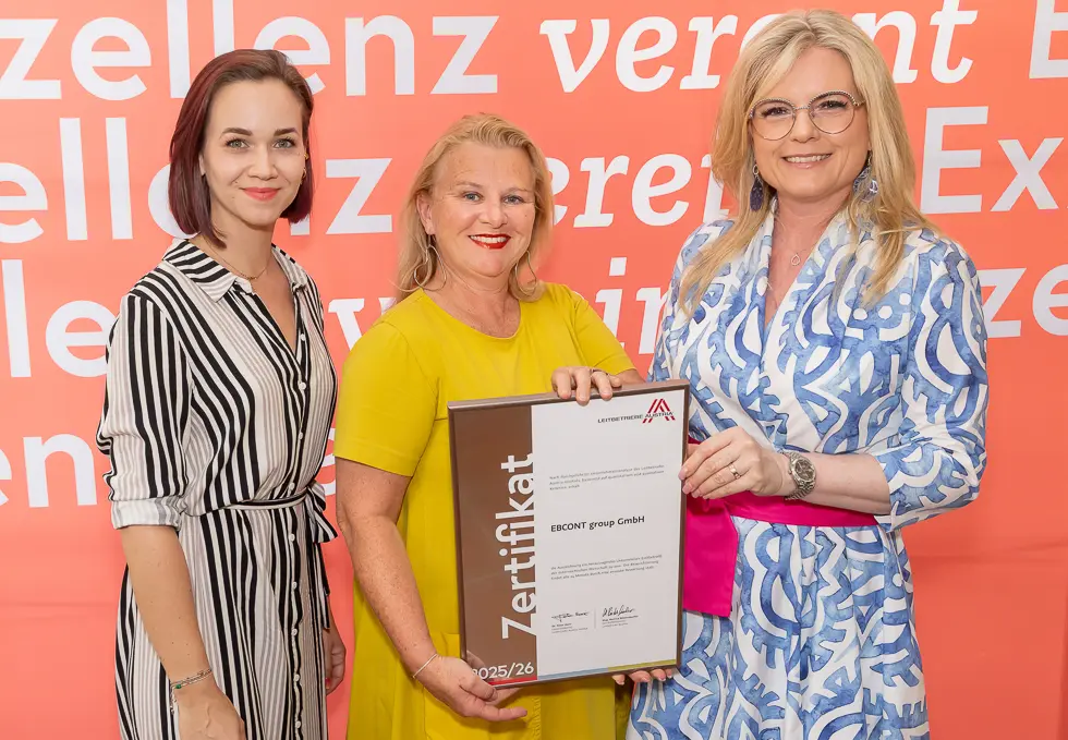 Ilse Merkinger-Boira, Bettina Richter, Monica Rintersbacher halten gemeinsam den Bilderrahmen mit dem neuen Zertifikat EBCONTs als Leitbetrieb Austria. Im Hintergrund eine orange Wand mit weißen Buchstaben. 
