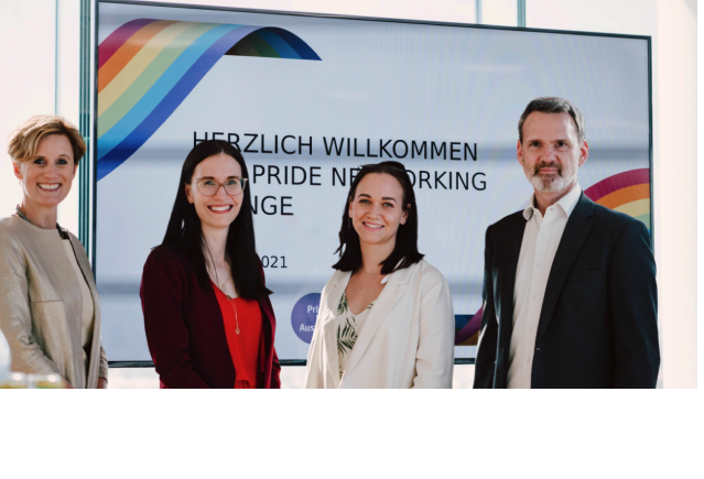Vier Personen - 2 Frauen und zwei Männer stehen gemeinsam vor einem großen Bildschirm mit einer Regenbogengrafik und der Aufschrift „Welcome PRIDE Networking Lounge“. Sie lächeln und sind business-casual gekleidet.