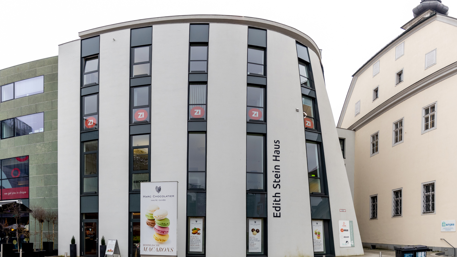 Moderne Gebäudefront des Edith Stein Hauses in Linz mit großen Glasfenstern und geschwungener Fassade. Im Erdgeschoss befindet sich ein Geschäft von Marc Chocolatier mit Werbeplakaten für Macarons. Links angrenzend ein grünes Gebäude, rechts ein historisches Bauwerk.