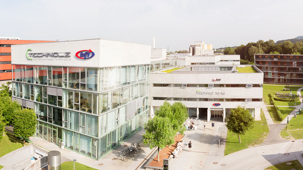 Modernes Bürogebäude im Technologiezentrum Techno-Z in Salzburg mit Glasfassade und begrüntem Flachdach. Auf dem Gebäude sind die Logos von Techno-Z und bfi sichtbar. Davor befinden sich Fahrräder, Bäume, ein kleiner Platz mit Sitzgelegenheiten sowie Personen im Außenbereich. Im Hintergrund weitere Büro- und Wohngebäude sowie grüne Parkflächen.