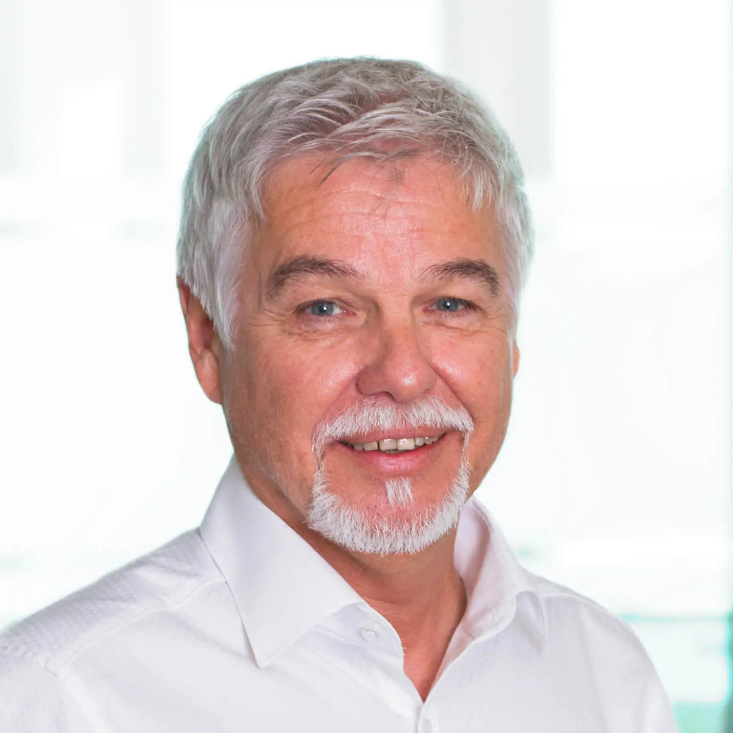 Älterer Mann mit weißem Haar und weiß-grauem Bart, trägt ein weißes Hemd und lächelt freundlich in die Kamera. Der Hintergrund ist hell und leicht unscharf, vermutlich eine moderne Innenraumumgebung.