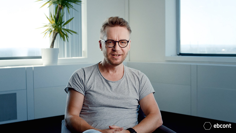 Ausschnitt aus dem Video auf dem Leopold Machacek entspannt in einem hellen Raum sitzt und in die Kamera lächelt. Er trägt ein hellgraues T-Shirt.