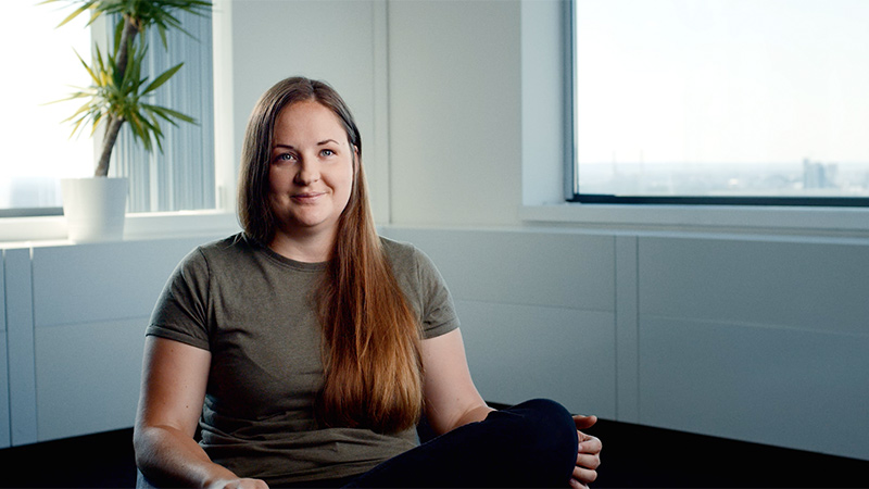 Ausschnitt aus dem Video auf dem Mitarbeiterin Carina entspannt in einem hellen Raum sitzt und lächelt in die Kamera. Sie trägt ein graues T-Shirt.