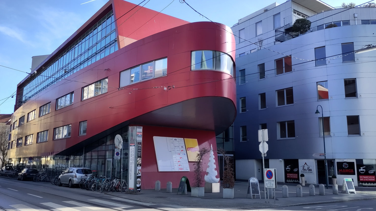 Modernes, rot-metallic glänzendes Bürogebäude in Graz mit markanter Rundung an der Ecke, Teil des Kommod-Hauses. Davor stehen Fahrräder, Autos und ein Aufsteller mit Lageplan. Rechts angrenzend ein silbernes Gebäude mit Gastronomiebetrieben im Erdgeschoss. Straßenschilder und Oberleitungen prägen das Stadtbild.