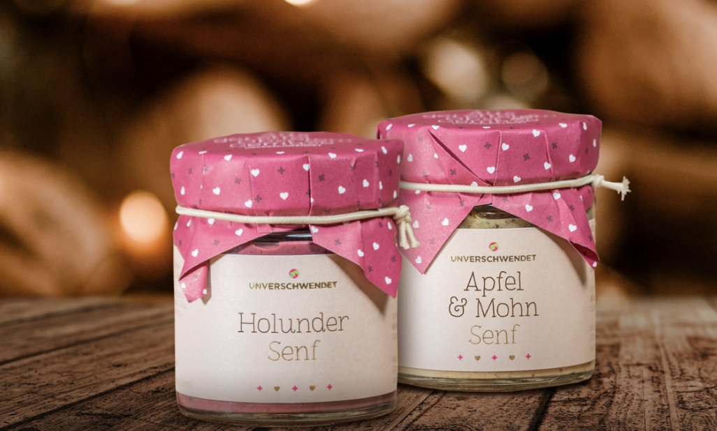 Zwei liebevoll verpackte Senfgläser der Marke UNVERSCHWENDET auf einem Holztisch – Sorten „Holunder Senf“ und „Apfel & Mohn Senf“.