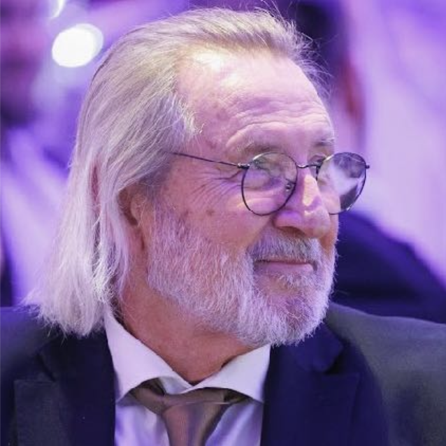 Älterer Mann mit grauem Haar und Bart, trägt eine runde Brille, ein dunkles Sakko und eine braune Krawatte. Er lächelt leicht und blickt nach rechts. Im Hintergrund sind unscharfe Personen und violettes Licht erkennbar.