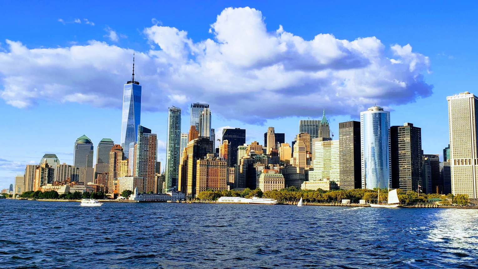 Panorama der Skyline von Manhattan, New York City, bei Tageslicht. Im Vordergrund ist der Hudson River mit Booten zu sehen, im Hintergrund ragen zahlreiche Hochhäuser und der markante One World Trade Center Tower in den blauen Himmel mit weißen Wolken.