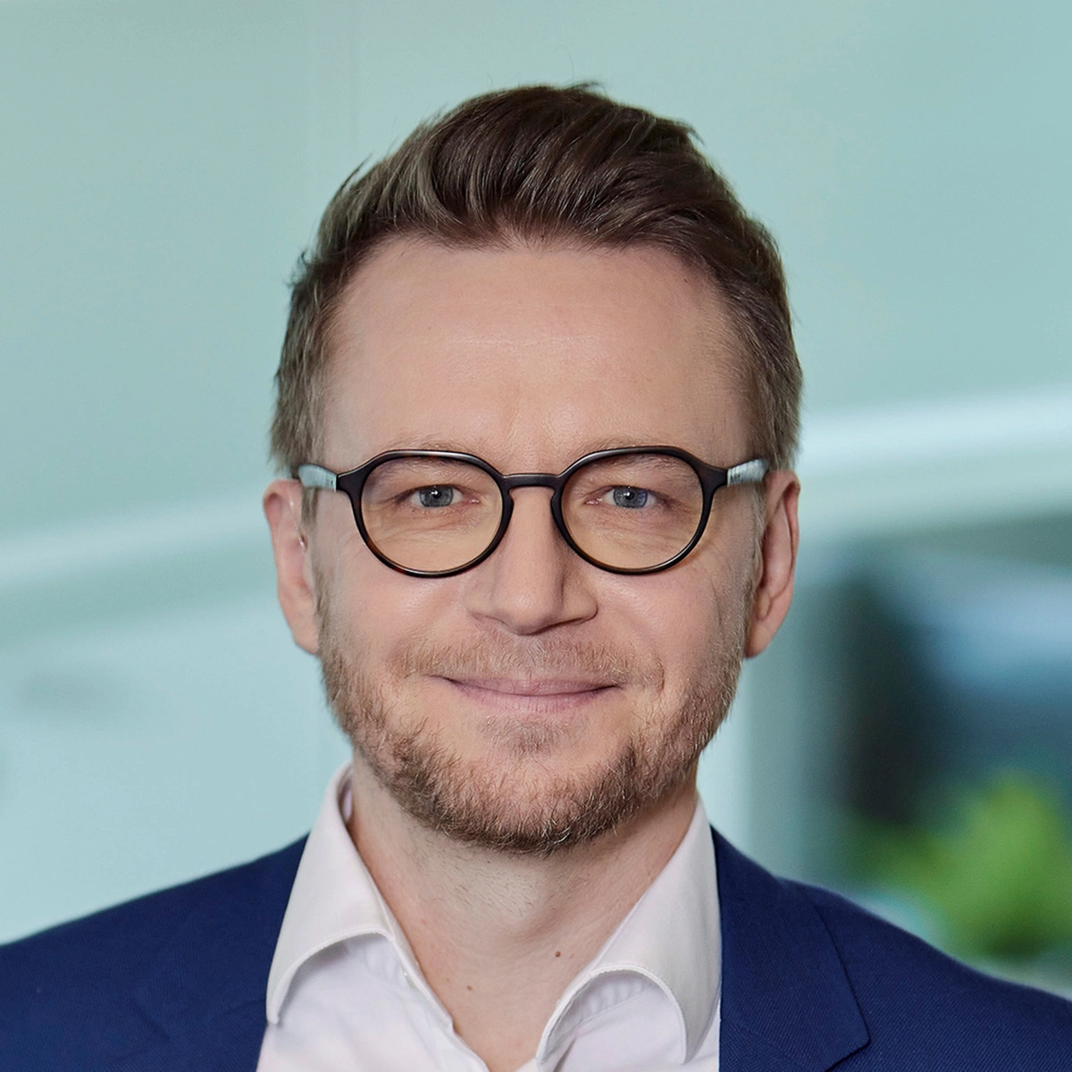 Mann mit kurz geschnittenem Haar, Bart und runder Brille, trägt ein weißes Hemd und ein blaues Sakko. Er lächelt leicht in die Kamera, im Hintergrund ist eine helle, moderne Büroumgebung zu erkennen.
