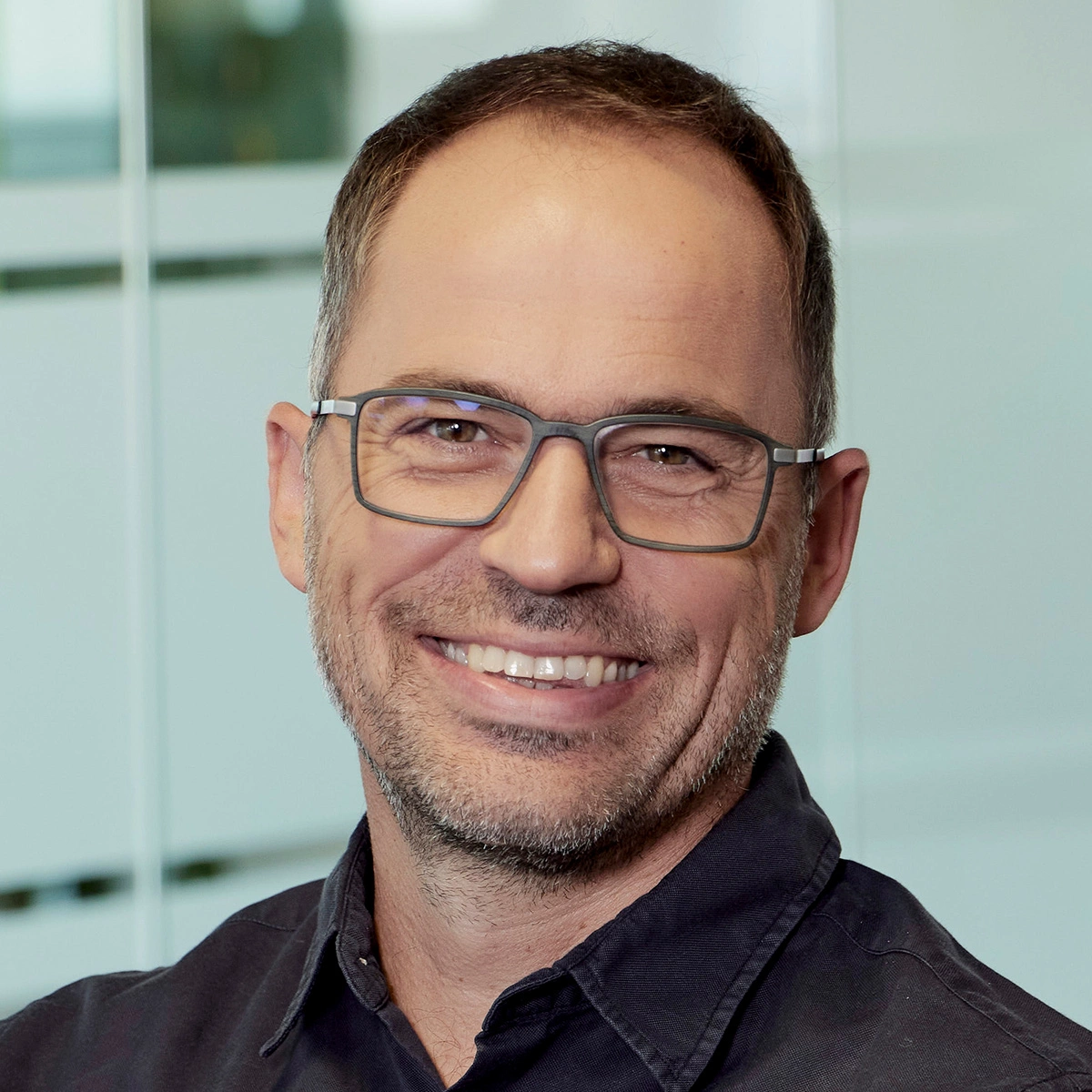 Mann mittleren Alters mit kurz geschnittenem Haar, Brille und Bart, trägt ein dunkles Hemd und lächelt freundlich in die Kamera. Im Hintergrund eine moderne, helle Büro- oder Glaswand.