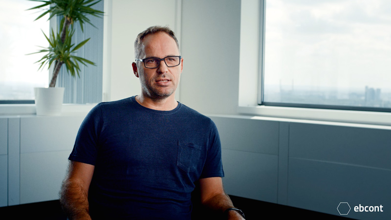 Ausschnitt aus dem Video auf dem Johannes Litschauer entspannt in einem hellem Raum sitzt. Er trägt ein dunkelblaues T-Shirt.