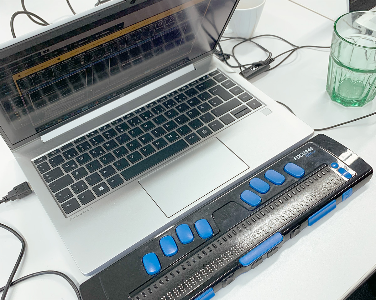 Laptop mit angeschlossener Braillezeile vom Typ Focus 40 Blue, auf einem Schreibtisch mit Kabeln, einem grünen Glas und einer weißen Tasse.