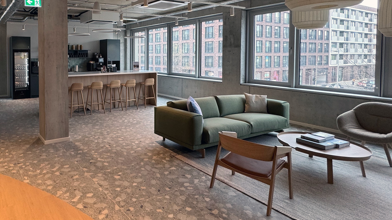 Moderner Büro-Loungebereich mit gemütlicher Sitzecke, grüner Couch, Holztischen und Bar mit hohen Hockern; große Fenster mit Blick auf ein urbanes Wohnviertel.