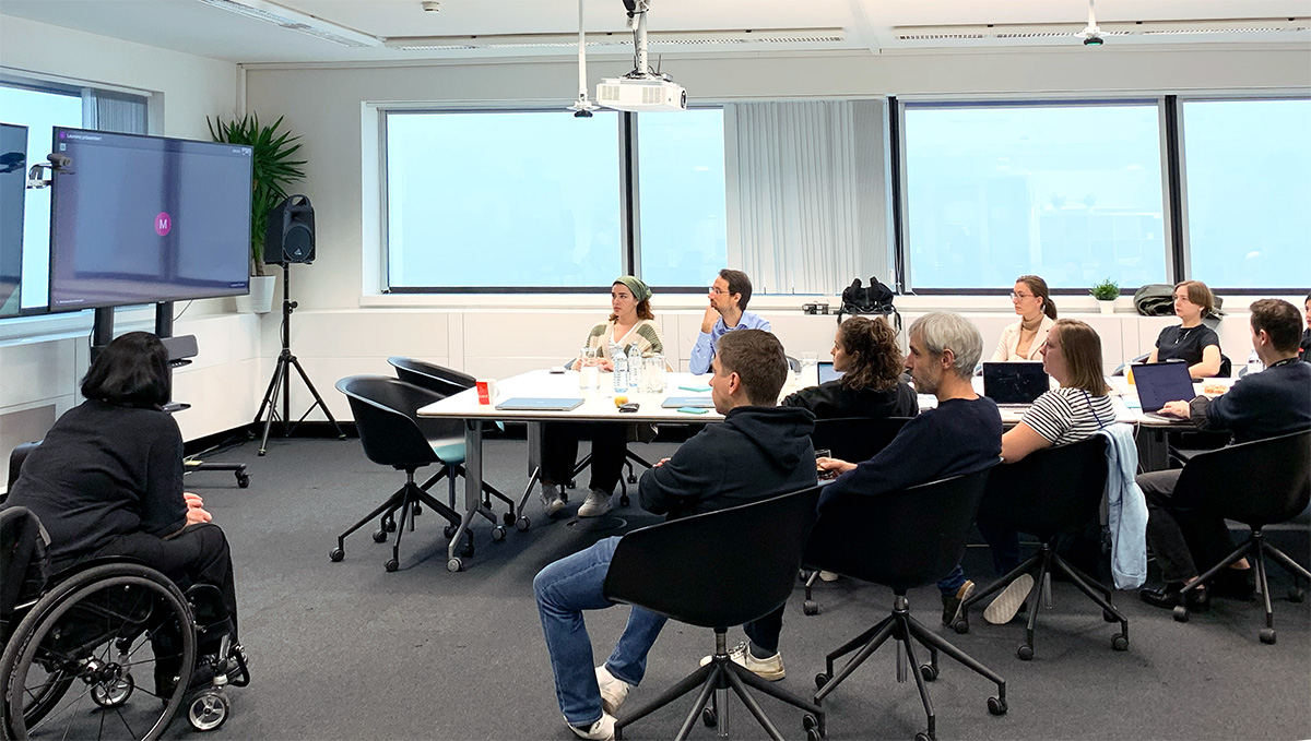 Eine Gruppe von Menschen sitzt in einem modernen Konferenzraum in einem Workshop. Eine Teilnehmerin im Rollstuhl ist ebenfalls anwesend. Alle blicken auf einen Bildschirm, auf dem eine virtuelle Teilnehmerin zugeschaltet ist. Auf dem Tisch stehen Laptops, Getränke und Notizblöcke.