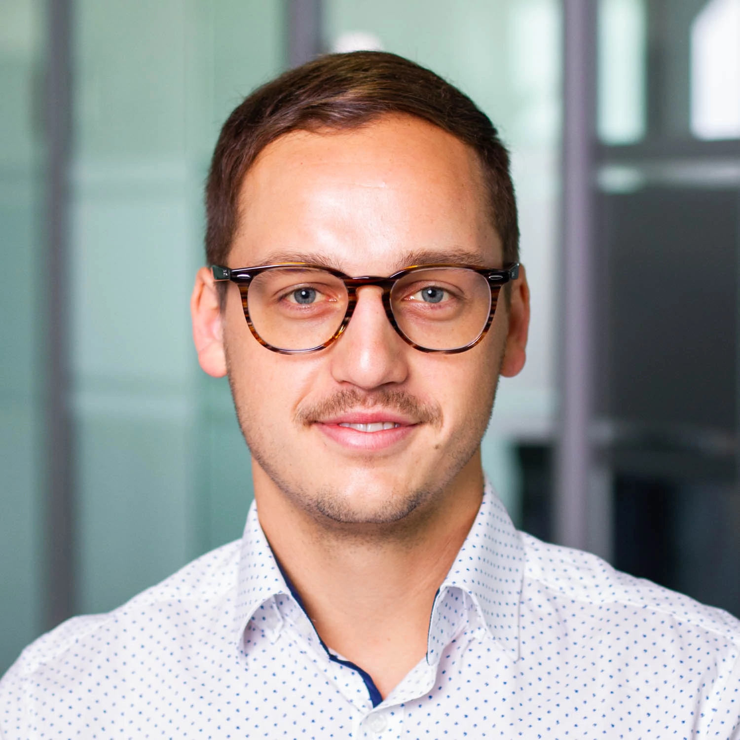 Junger Mann mit braunen Haaren, Brille und Oberlippenbart, trägt ein weißes Hemd mit feinem Muster und blickt freundlich in die Kamera. Im Hintergrund ist eine moderne Büroeinrichtung mit Glastrennwänden zu erkennen.