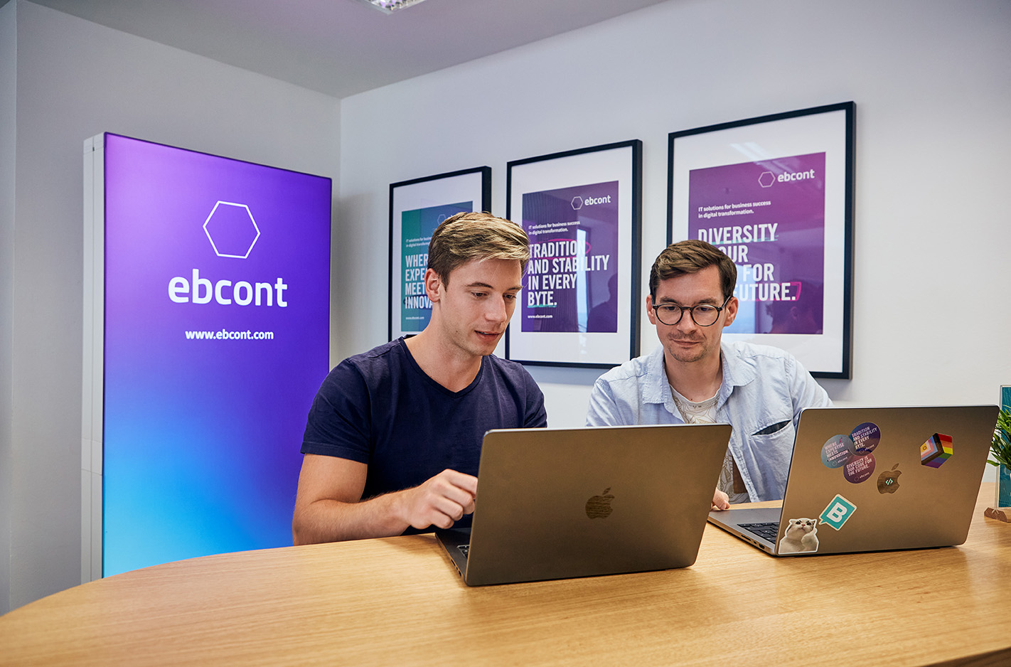Zwei junge Männer sitzen gemeinsam an einem Tisch in einem EBCONT-Besprechungsraum und arbeiten an ihren Laptops. Im Hintergrund ist ein beleuchtetes EBCONT-Logo zu sehen. Auf einem Laptop sind Pride- und Tech-Sticker angebracht.