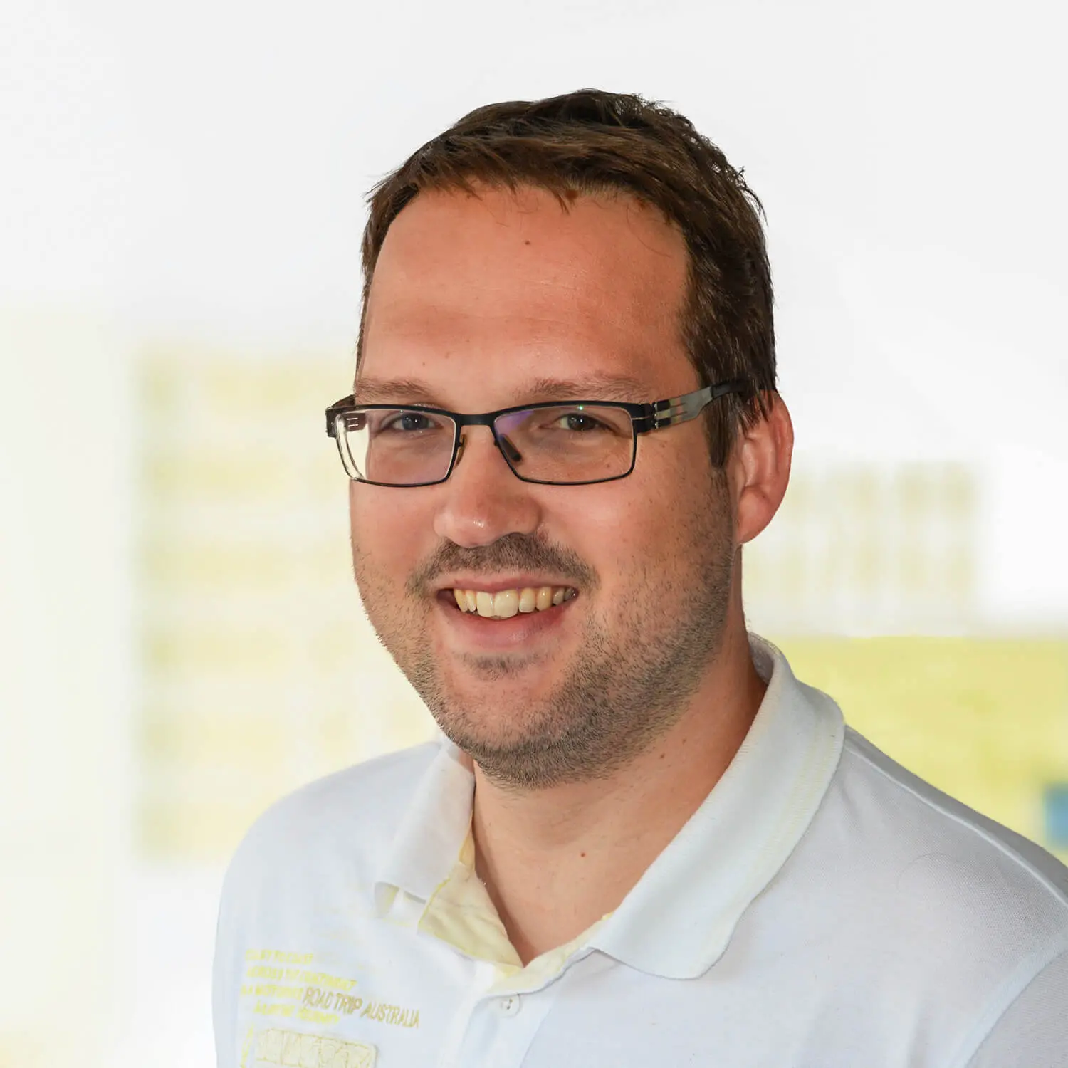 Mann mit kurzen braunen Haaren, Brille und leichtem Bartansatz, trägt ein weißes Poloshirt mit gelbem Aufdruck und lächelt in die Kamera. Der Hintergrund ist hell und unscharf.