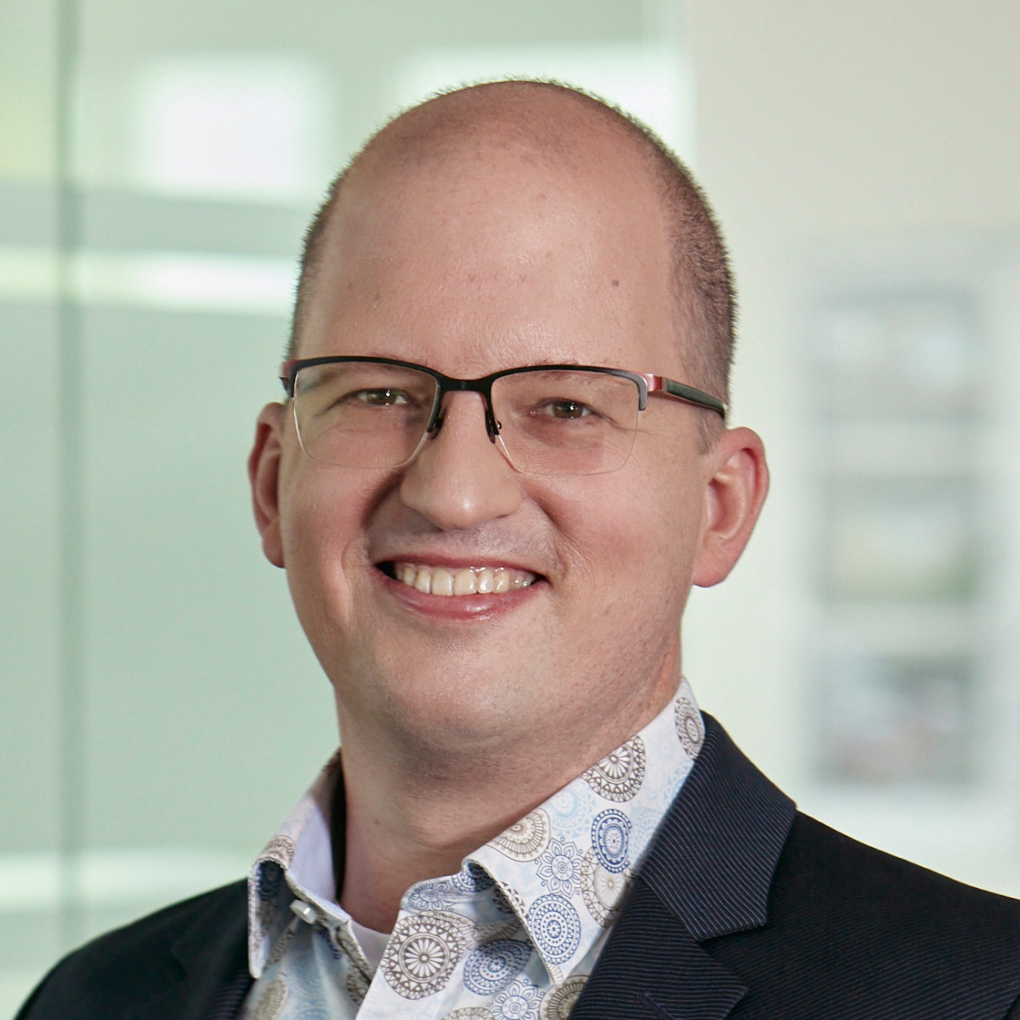 Mann mit Glatze und Brille, trägt ein dunkles Sakko und ein gemustertes Hemd. Er lächelt in die Kamera, im Hintergrund ist eine helle, moderne Büroumgebung zu erkennen.