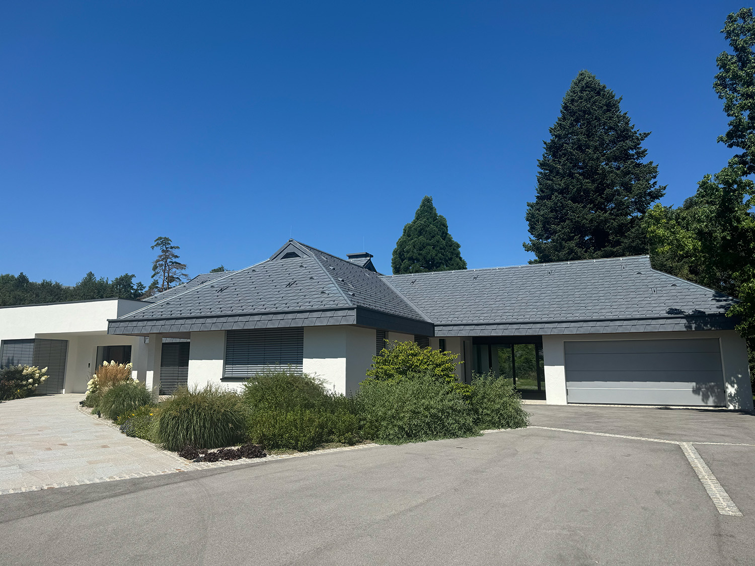  Modernes, einstöckiges Wohnhaus mit dunkelgrauem Walmdach, weißer Fassade, großen Fenstern und gepflegtem Vorgarten, aufgenommen bei strahlend blauem Himmel.