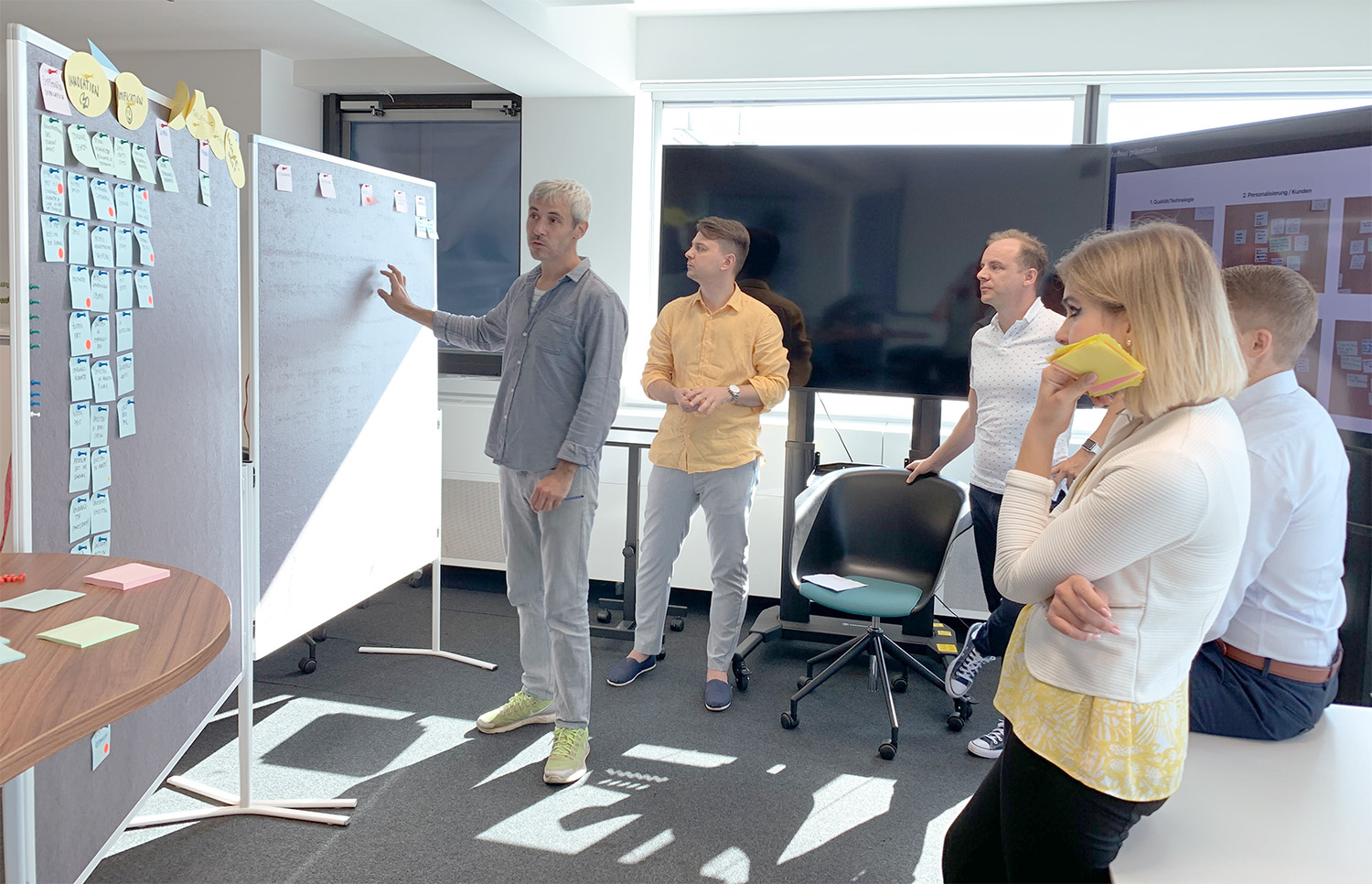Fünf Personen stehen in einem modernen Büro um eine beschriebene Pinnwand, die mit vielen Haftnotizen versehen ist. Eine Person erklärt, während die anderen aufmerksam zuhören. Auf dem Tisch liegen weitere bunte Notizzettel.