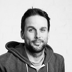 Porträt eines lächelnden Mannes mit Bart in einem Kapuzenpullover vor hellem Hintergrund, in Schwarz-Weiß aufgenommen.