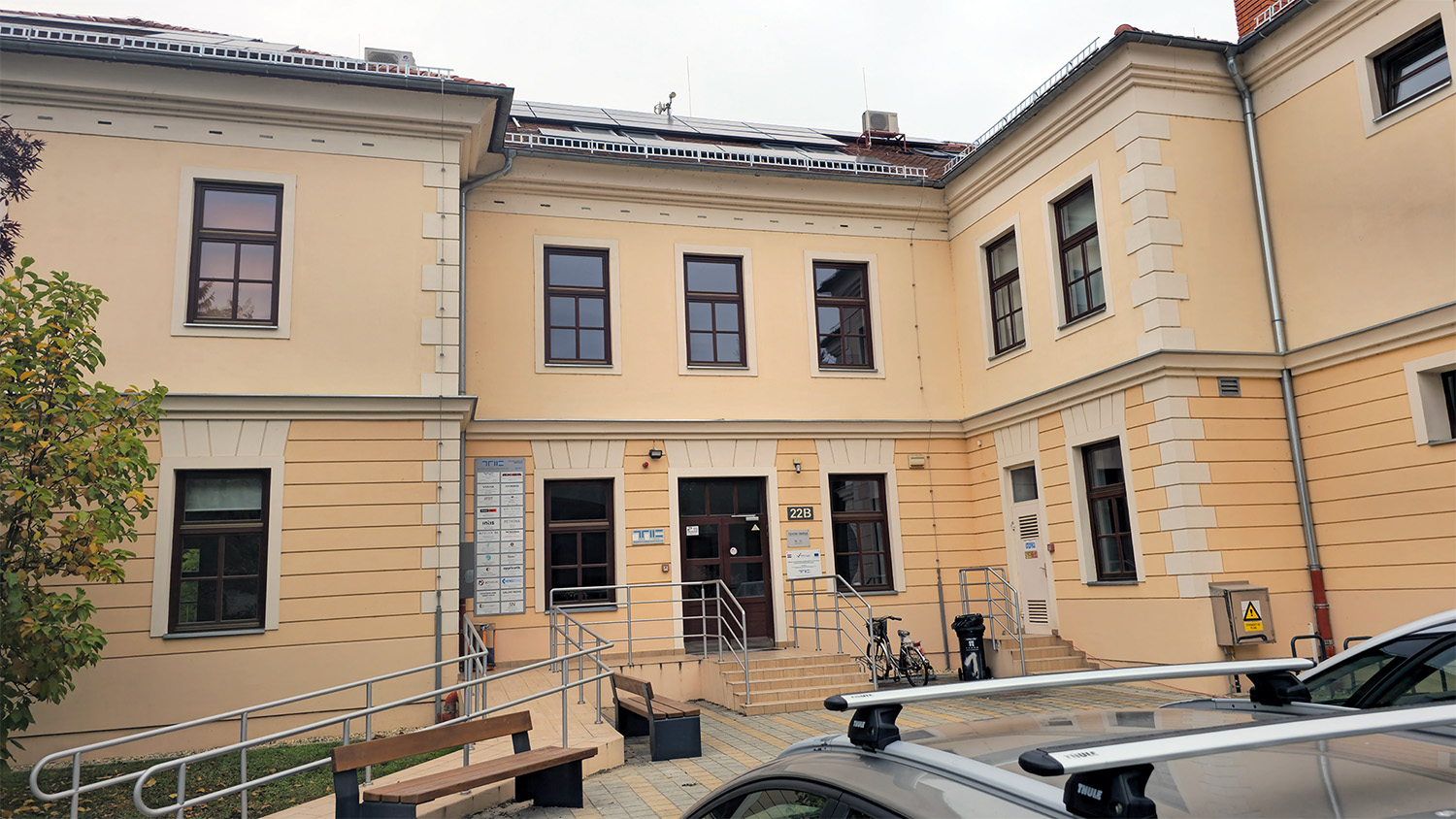 Zweigeschossiges, hellgelbes Bürogebäude mit braunen Fensterrahmen und Eingangsbereich mit Rampe und Treppe. Vor dem Gebäude stehen Fahrräder, Bänke und ein geparktes Auto.