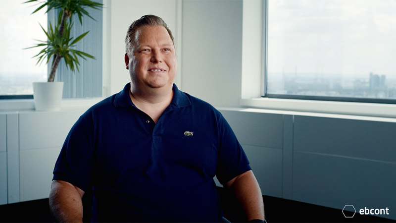 Ausschnitt aus dem Video auf dem Alexander Deles entspannt in einem hellen Raum sitzt und in die Kamera lächelt. Er trägt ein dunkelblaues Polo T-Shirt.