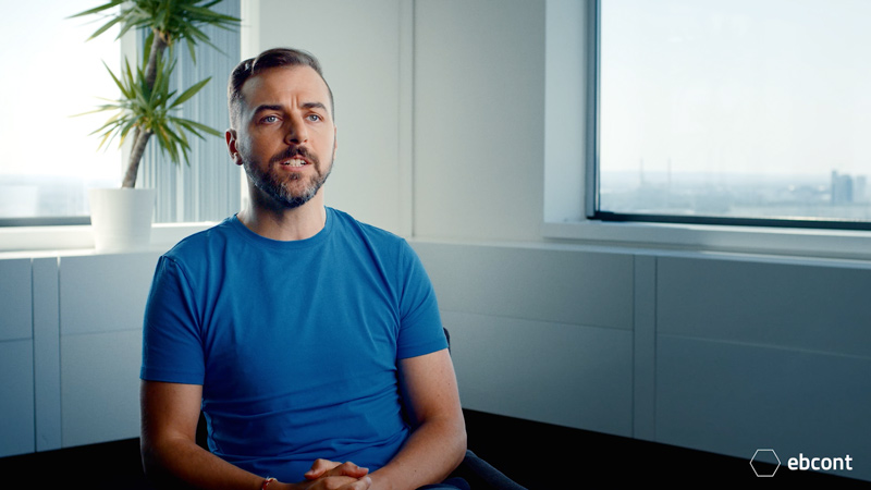 Ausschnitt aus dem Video auf dem Mitarbeiter Mathias entspannt in einem hellen Raum sitzt. Er trägt ein blaues T-Shirt und Brille.