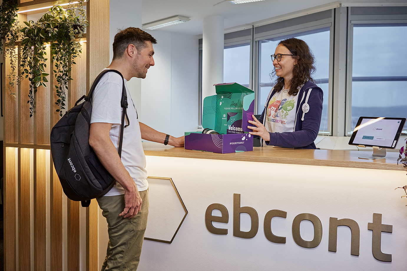 Ein neuer Mitarbeiter wird an der EBCONT-Rezeption herzlich mit einer „Welcome-Box“ begrüßt. Er trägt einen Rucksack mit EBCONT-Logo, während eine Kollegin ihm freundlich das Willkommenspaket überreicht.