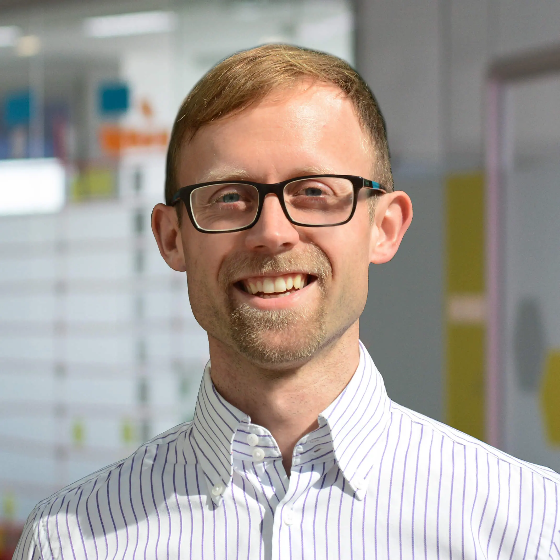 Mann mit kurzem, blondem Haar, Brille und Bartansatz, trägt ein weißes Hemd mit feinen blauen Streifen und lächelt freundlich in die Kamera. Im Hintergrund ist eine moderne Büro- oder Arbeitsumgebung mit Glaswänden und farbigen Elementen zu erkennen.