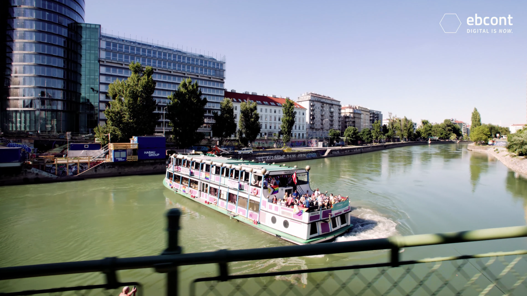 Das festlich geschmückte Schiff „Vindobona“ fährt mit vielen Menschen an Bord beim EBCONT Pride-Boot Event den Donaukanal in Wien entlang. Am Boot wehen Regenbogenfahnen, am Ufer sind moderne und historische Gebäude sowie Street-Art zu sehen.