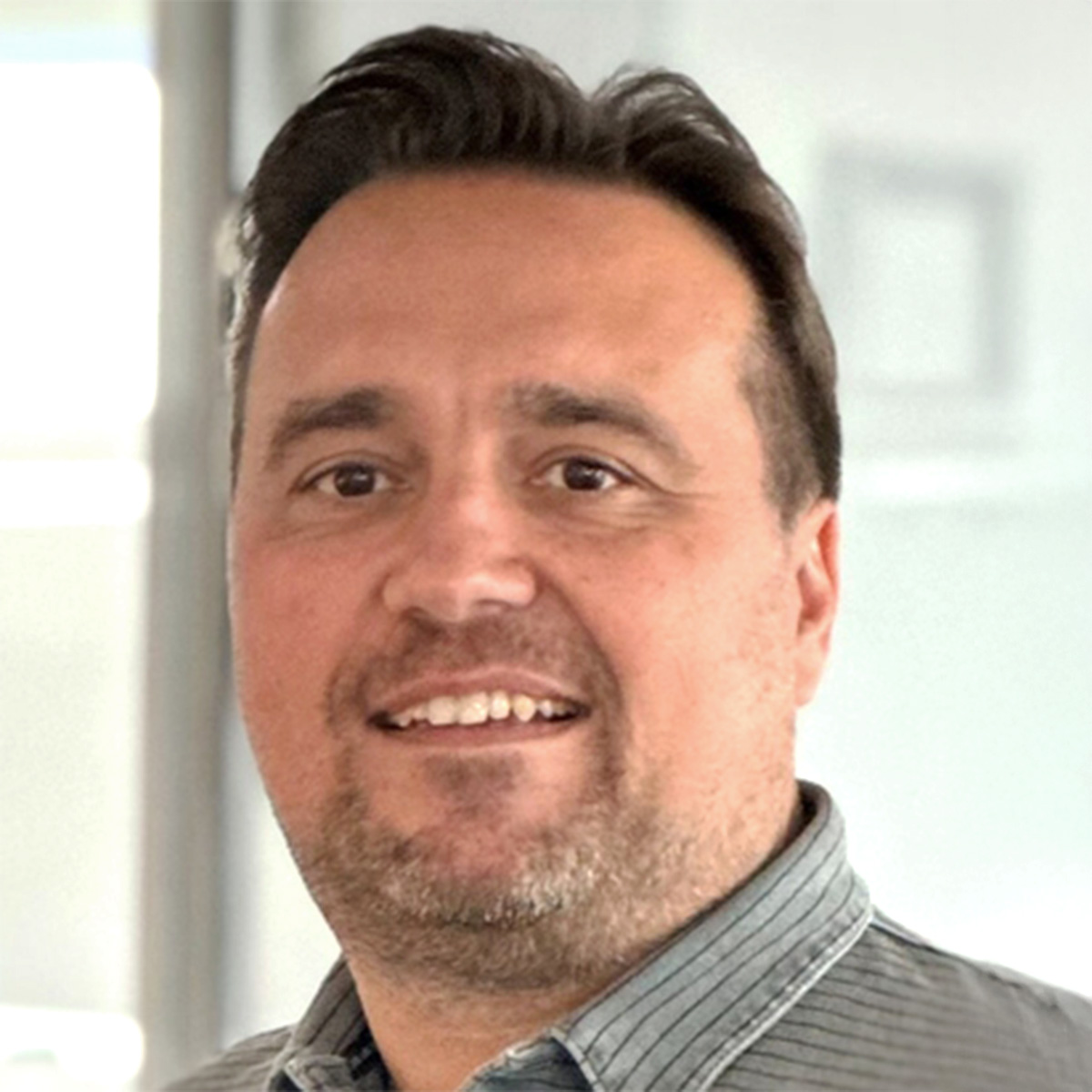 Porträt eines lächelnden Mannes mit kurzem dunklem Haar und Bartstoppeln, gekleidet in ein graues Hemd mit feinem Muster, vor einem hellen, unscharfen Bürohintergrund.