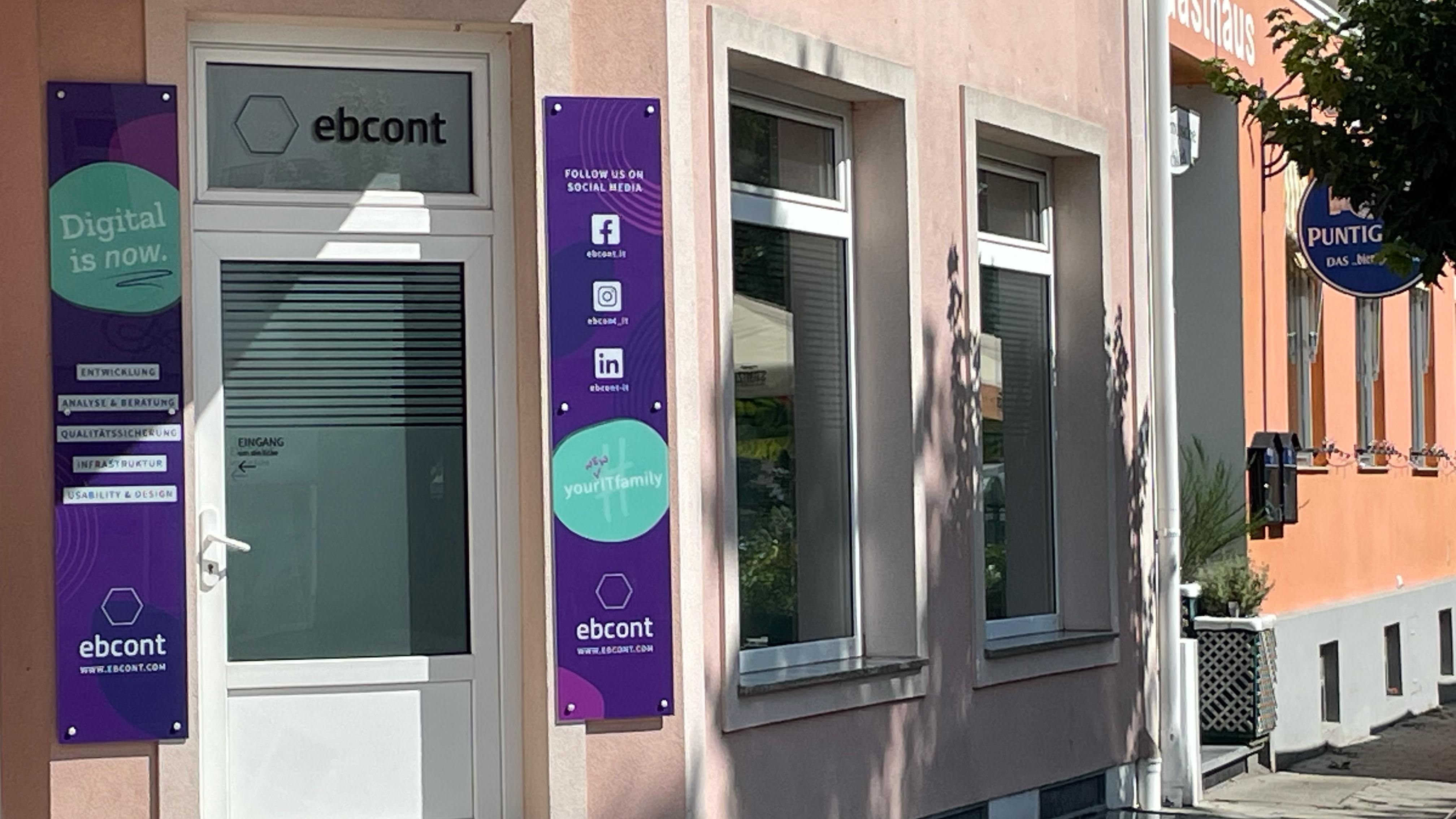 Eingangsbereich einer EBCONT-Niederlassung mit heller Fassade und violetten Firmenschildern links und rechts der Tür. Auf den Tafeln sind Slogans wie „Digital is now.“, Leistungsbereiche wie „Entwicklung“ und „Qualitätssicherung“ sowie Social-Media-Kanäle zu sehen. Rechts im Bild befindet sich ein angrenzendes Gebäude mit einem Bierschild.